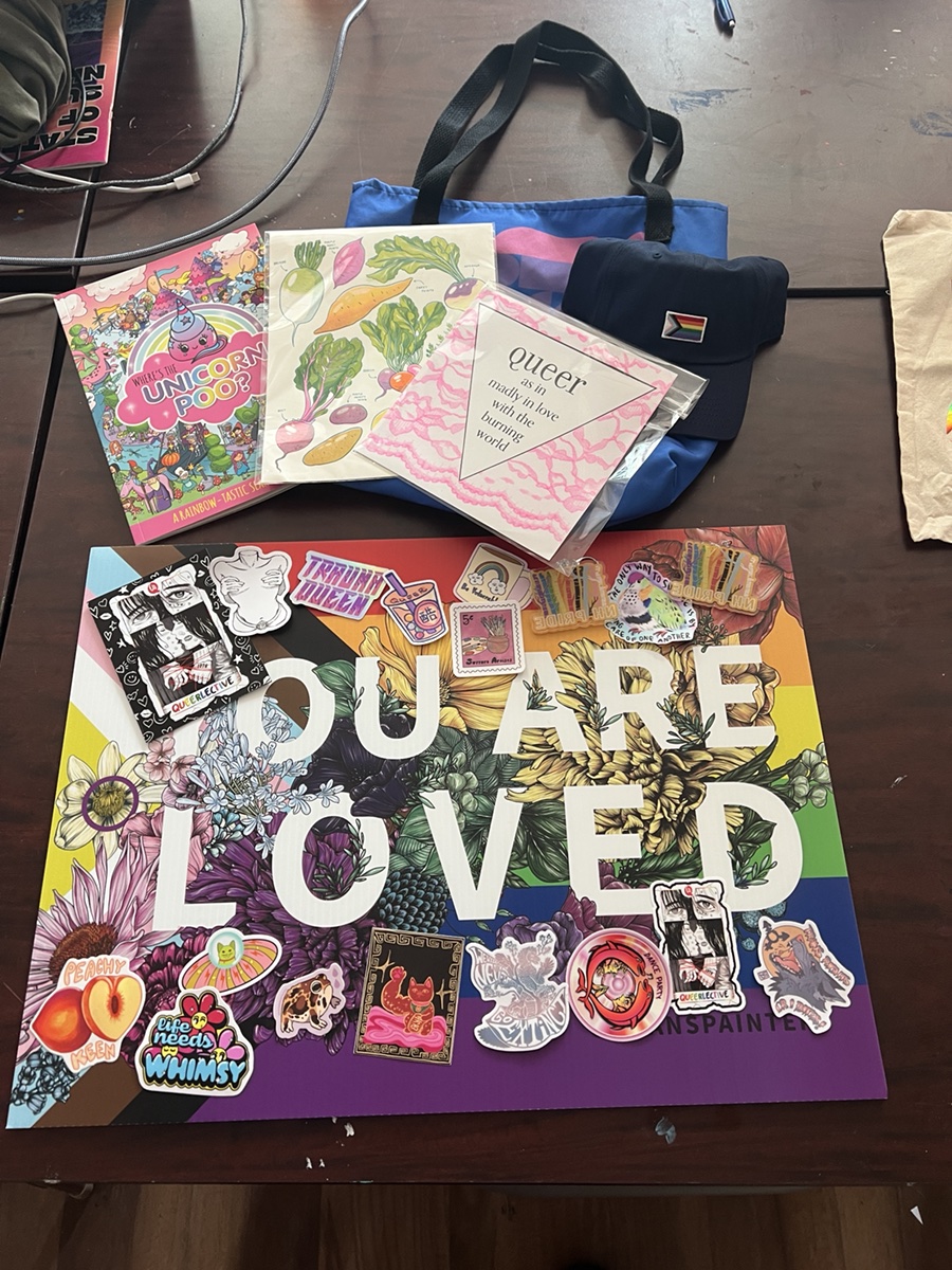 Raffle Basket 1 with a blue Queerlective tote, cap, art, book, and stickers